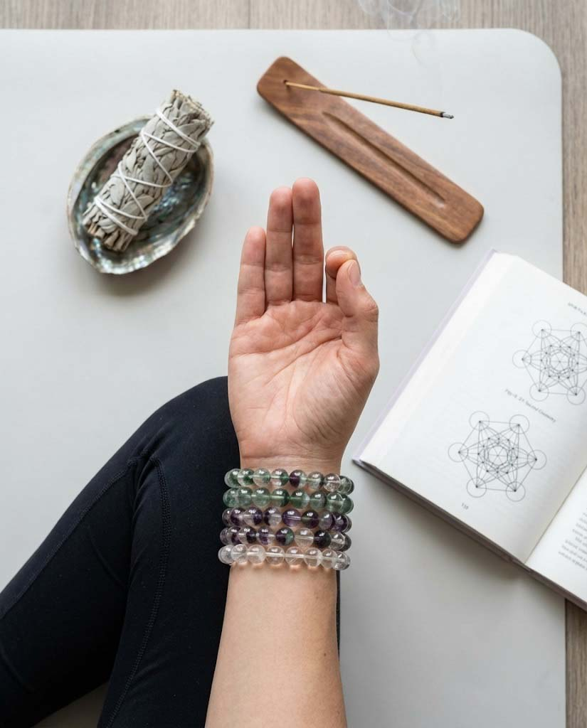 Hand in a meditation mudra gesture on a clean yoga mat, wearing a stack of green and purple fluorite crystal bracelets next to white sage and incense.