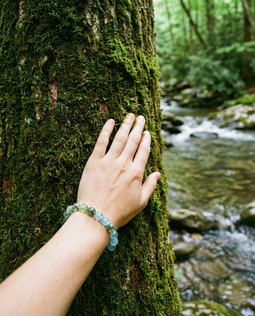 Hand touching a mossy tree trunk in a forest, wearing natural green and blue aquamarine gemstone bracelets that harmonize with the nature background.